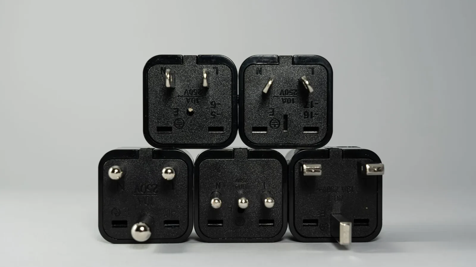 Six travel adapters arranged on a white surface showing Type C, G, I and universal plug configurations with USB ports visible