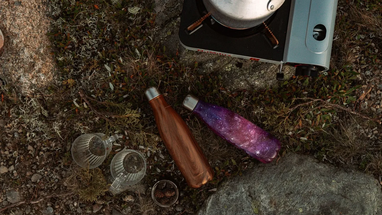Stainless steel and recycled-plastic travel water bottles lined up against a worn hiking backpack showing size comparison