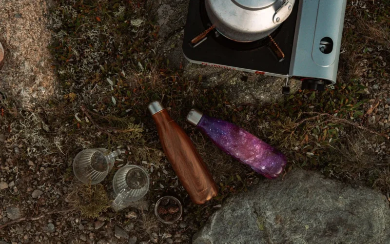 Stainless steel and recycled-plastic travel water bottles lined up against a worn hiking backpack showing size comparison